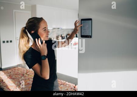 Ragazza adolescente che parla su smartphone mentre si utilizza un tablet digitale montato su una parete bianca a casa Foto Stock