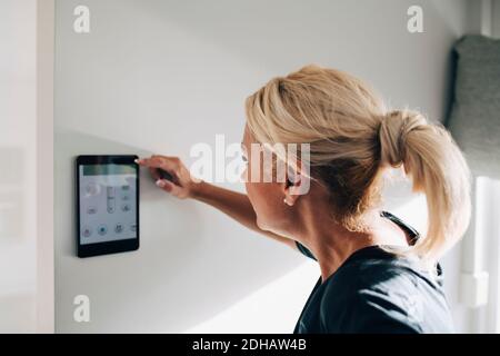 Vista laterale del termostato biondo per la regolazione della donna tramite tablet digitale montato su una parete bianca a casa Foto Stock