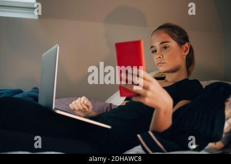 Ragazza adolescente che prende selfie dallo smartphone mentre si sdraia con computer portatile a casa Foto Stock