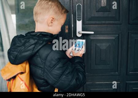 Vista posteriore del ragazzo utilizzando l'app sullo smartphone a. sbloccare la porta della casa Foto Stock
