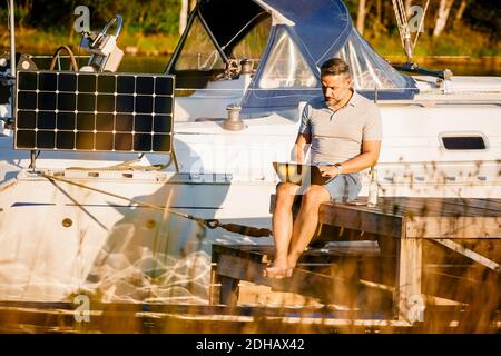 Uomo maturo che lavora su un computer portatile mentre si siede al molo contro yacht sul lago Foto Stock