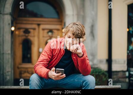 Ragazzo adolescente che usa il telefono cellulare mentre si siede sulla panca dentro città Foto Stock