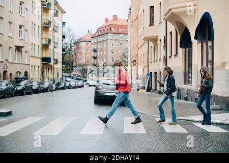 Tutta la lunghezza degli amici che usano il telefono cellulare mentre attraversano la strada in città Foto Stock