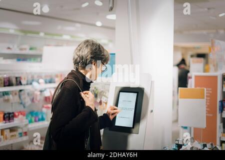 Vista laterale della donna anziana che usa il chiosco presso il negozio di farmacia Foto Stock