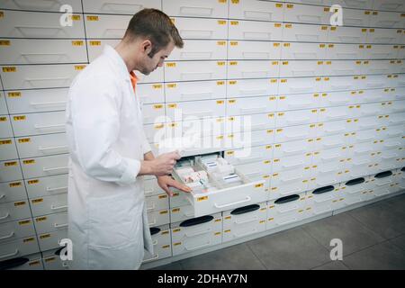 Vista laterale del farmacista maschile maturo che tiene in piedi la carta da prescrizione da farmaci in cassetto in negozio Foto Stock