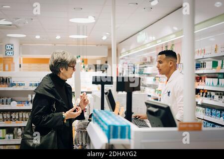 Vista laterale di una donna senior che parla con il farmacista maschile al momento del check-out in negozio Foto Stock