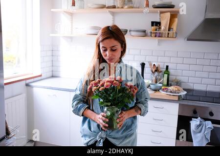 Donna che porta vaso di fiori in piedi in cucina Foto Stock