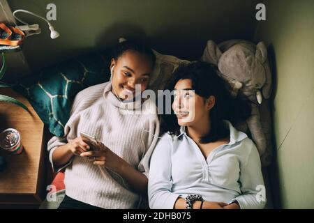 Direttamente sopra il colpo delle ragazze sorridenti che ascoltano la musica sopra telefono cellulare mentre si sdraiano sul letto a casa Foto Stock