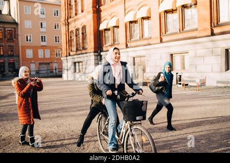 Giovani donne che fotografano la ragazza adolescente in bicicletta con l'amico contro la costruzione in città Foto Stock