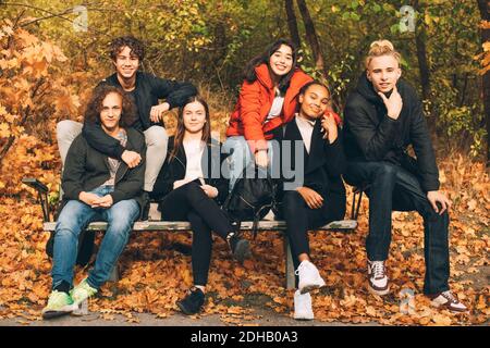 Ritratto completo di amici adolescenti sorridenti seduti insieme panca del parco durante l'autunno Foto Stock