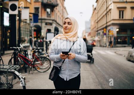 Giovane donna musulmana che guarda via tenendo uno smartphone mentre cammina sulla strada in città Foto Stock