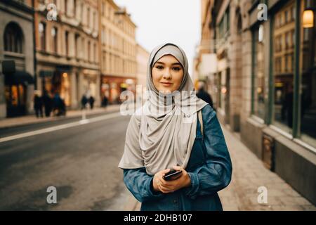 Ritratto di giovane donna sicura che indossa hijab in piedi con il cellulare telefono sul marciapiede in città Foto Stock