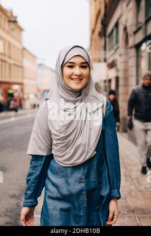 Ritratto di giovane donna musulmana sorridente che indossa hijab camminando sopra marciapiede in città Foto Stock