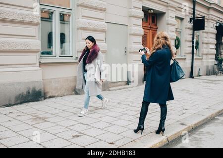 Vista posteriore di una donna che fotografa un amico che cammina sul marciapiede contro la costruzione in città Foto Stock