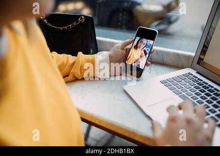 La sezione centrale della freelance femminile che tiene lo smartphone usando il laptop a. caffè Foto Stock