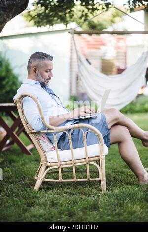 Vista laterale di un uomo maturo che lavora su un computer portatile mentre è seduto su sedia in cortile Foto Stock