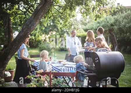 Ragazzo che prepara il cibo alla griglia con la madre mentre la famiglia godendo al tavolo da pranzo in festa cortile Foto Stock