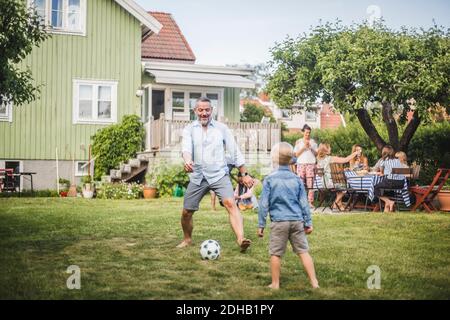 Padre che gioca a calcio con il figlio mentre gli amici si divertono a. tavolo in festa cortile Foto Stock