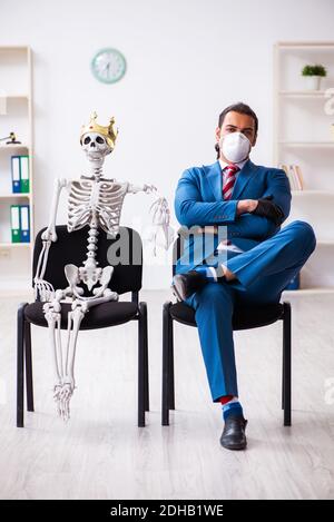 Giovane uomo d'affari e scheletro in ufficio in concetto pandemico Foto Stock