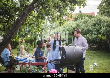 Uomo che parla con la donna con il figlio che prepara il cibo al barbecue grigliate contro gli amici durante la festa del fine settimana Foto Stock