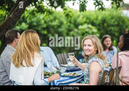 Donna sorridente che tiene un bicchiere di vino mentre si siede con gli amici a. festa in giardino Foto Stock