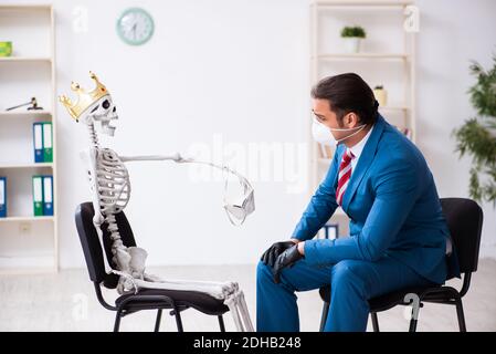 Giovane uomo d'affari e scheletro in ufficio in concetto pandemico Foto Stock