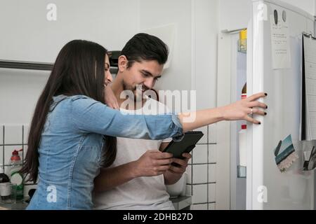 Sorridente coppia che usa il tablet digitale mentre si è in piedi in frigorifero a. cucina Foto Stock