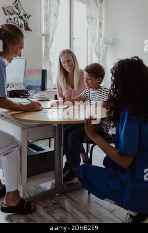 Medico che guarda il paziente che punta sulla carta mentre l'infermiera femminile e madre seduta alla scrivania in ospedale Foto Stock