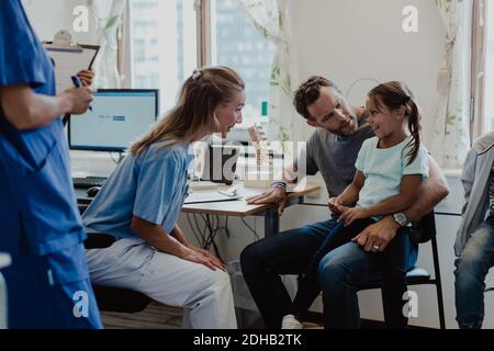 Donna pediatra che parla con una ragazza sorridente seduta con il padre dentro sala di visita medica Foto Stock