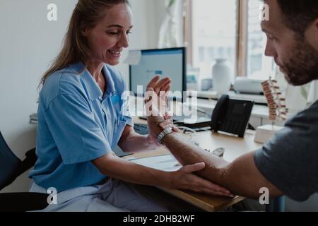 Sorridente medico che esamina la mano del paziente maschio mentre si siede in medicina sala esame in clinica Foto Stock