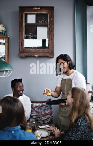 Sorridente proprietario che serve cibo a giovane uomo e donne a. tavolo da pranzo Foto Stock