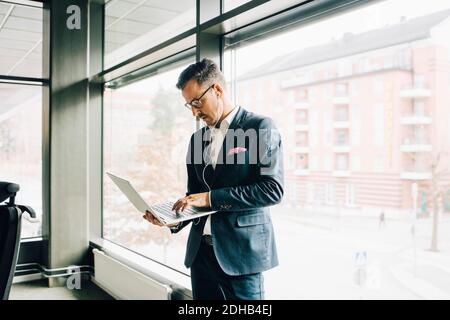 Uomo d'affari maturo che usa il computer portatile mentre si trova contro la finestra in ufficio Foto Stock