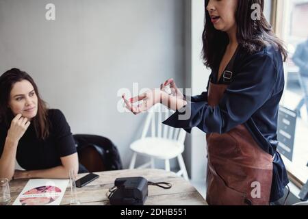 Sezione centrale del profumo di spruzzatura della donna sul polso da una collega femminile seduto al tavolo in officina Foto Stock