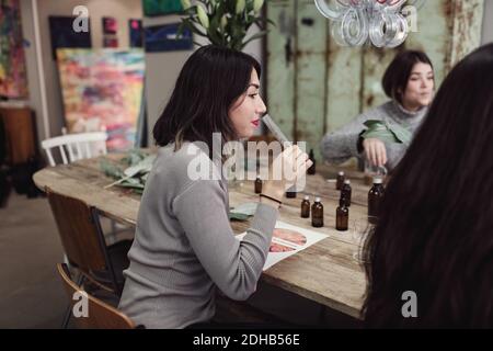 Vista laterale della giovane donna che puzzava di liquido dalla provetta mentre si siede con i colleghi al tavolo in officina Foto Stock
