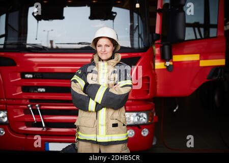 Ritratto di sicuro pompiere femminile in piedi con le braccia incrociate motore antincendio alla stazione dei vigili del fuoco Foto Stock