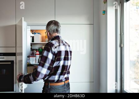 Vista posteriore dell'uomo anziano in pensione in piedi vicino al frigorifero aperto porta in cucina a casa Foto Stock