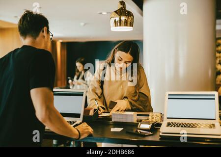 Uomo che guarda una donna che firma alla reception dell'hotel lobby Foto Stock