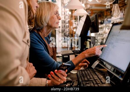 Cliente femminile senior e venditore che discutono sopra il monitor del computer a. checkout in negozio di hardware Foto Stock