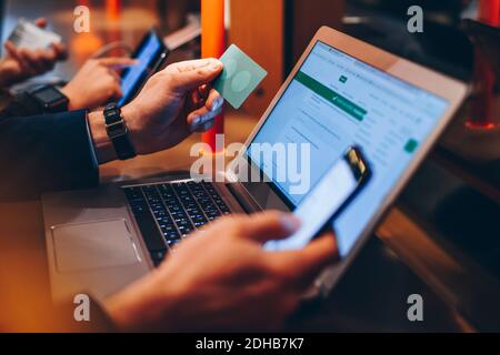 Immagine ritagliata di un uomo d'affari che detiene la carta di credito mentre si utilizza il cellulare telefono e computer portatile in aeroporto Foto Stock