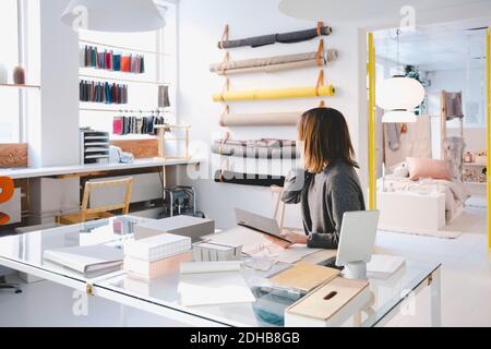 Donna imprenditore che guarda i campioni di tessuto mentre tiene il cuscinetto della nota al tavolo in negozio Foto Stock