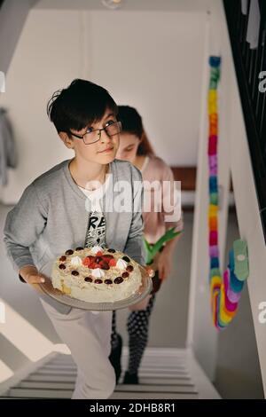 Ragazzo che porta una torta di compleanno mentre salendo i gradini con la sorella a casa Foto Stock