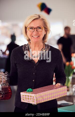 Ritratto di donna anziana sorridente che tiene scatola di regalo e succo mentre si sta a casa Foto Stock