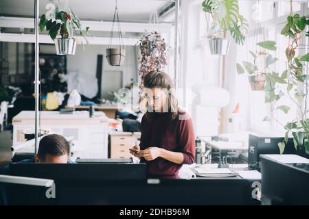 Donna d'affari che usa il telefono cellulare mentre si trova in piedi da un collega maschio ufficio creativo Foto Stock
