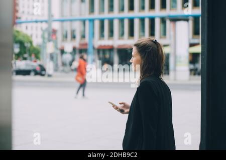 Vista laterale di una donna d'affari sicura che tiene il telefono cellulare mentre cammina sulla strada in città Foto Stock