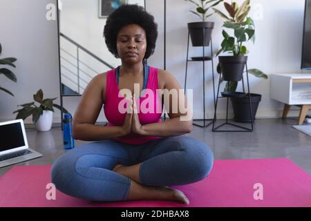 Donna afro-americana meditating seduta su tappetino indossando abiti sportivi. Laptop sullo sfondo. Auto isolamento fitness benessere tecnologia a casa Foto Stock