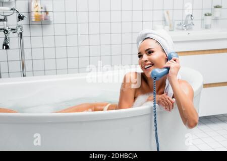 Donna allegra in asciugamano sulla testa che parla al telefono in vasca da bagno con schiuma Foto Stock