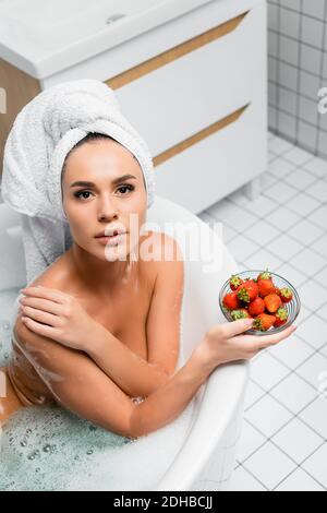 Giovane donna con asciugamano sulla testa guardando la macchina fotografica mentre tenendo il recipiente delle fragole nella vasca da bagno Foto Stock