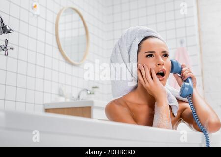 Donna scioccata in asciugamano che parla al telefono mentre si siede vasca da bagno a casa Foto Stock