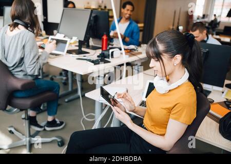 Hacker di computer IT femminile che utilizzano tablet digitale mentre i colleghi lavorano in ufficio creativo Foto Stock