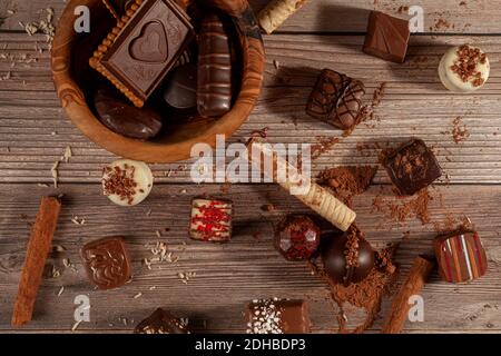 Una ciotola di cioccolatini assortiti e biscotti al cioccolato su sfondo di legno con bastoncini di cacao in polvere alla cannella, pirouettes e cocco spruzzano la creazione Foto Stock